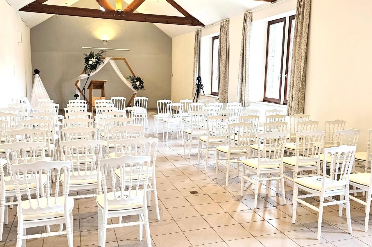 Salle lumineuse aménagée pour une cérémonie, avec des chaises blanches alignées face à une arche décorée de fleurs.