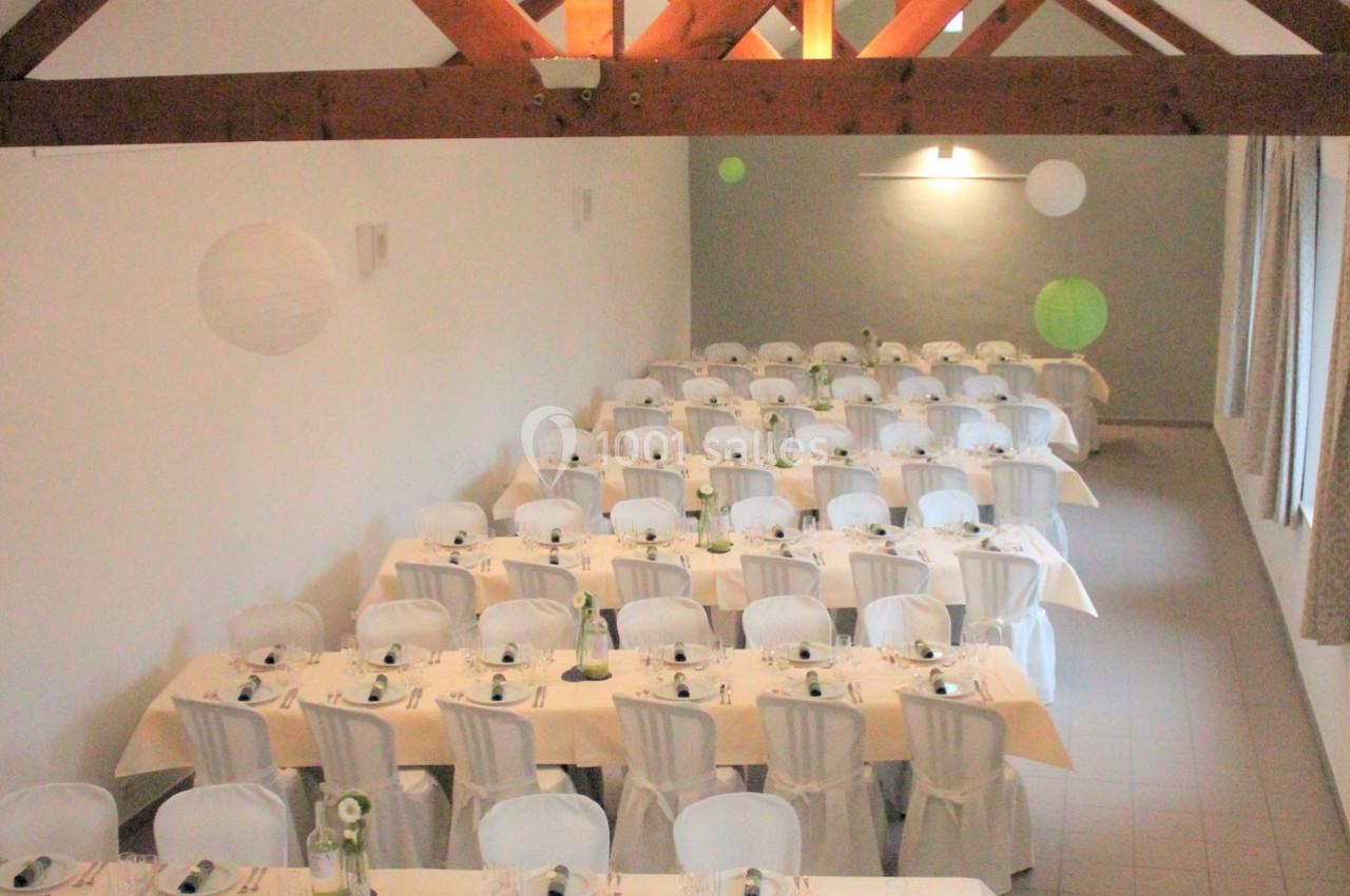 Salle de réception avec des tables alignées, nappées de blanc, décorées de vaisselle et lanternes suspendues.