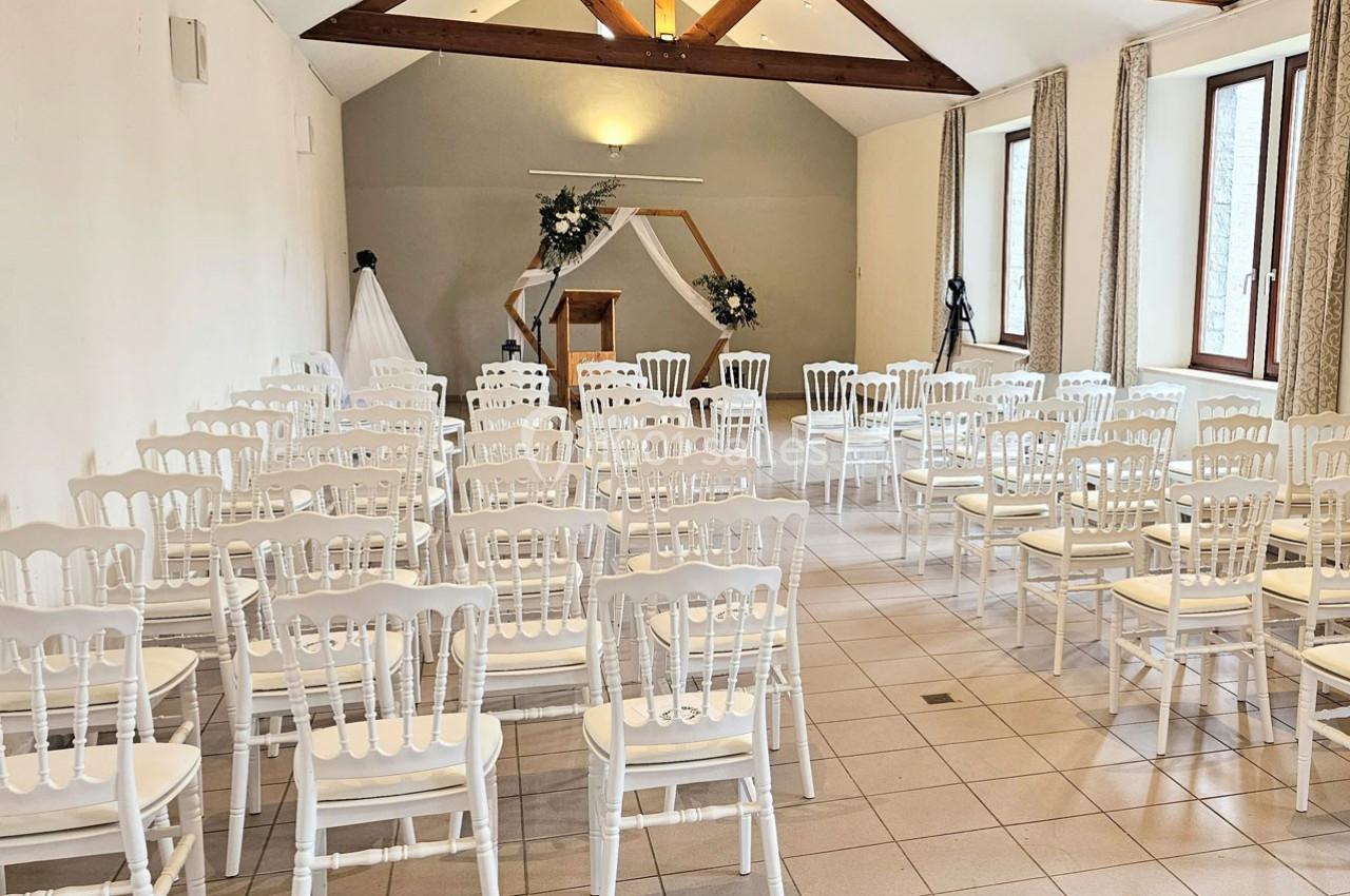 Salle lumineuse aménagée pour une cérémonie avec des chaises blanches alignées et une arche décorée de fleurs.