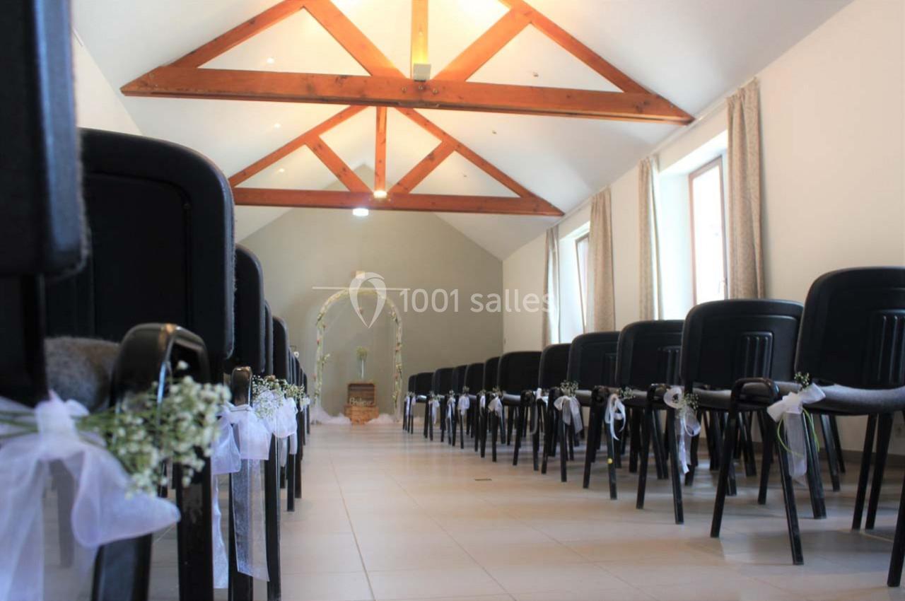 Salle lumineuse aménagée pour une cérémonie, avec des chaises décorées de rubans blancs et une arche florale au fond.