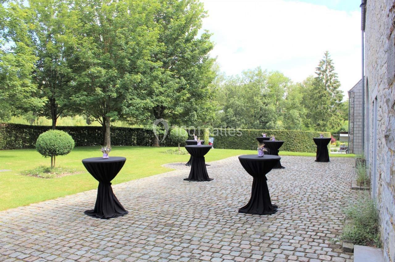 Tables hautes noires dressées sur une terrasse pavée entourée de verdure et d'arbres.