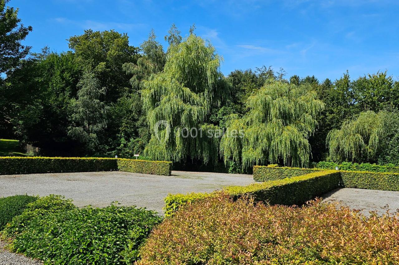 Carré de gravier entouré de haies taillées, avec des saules pleureurs et une végétation dense en arrière-plan.