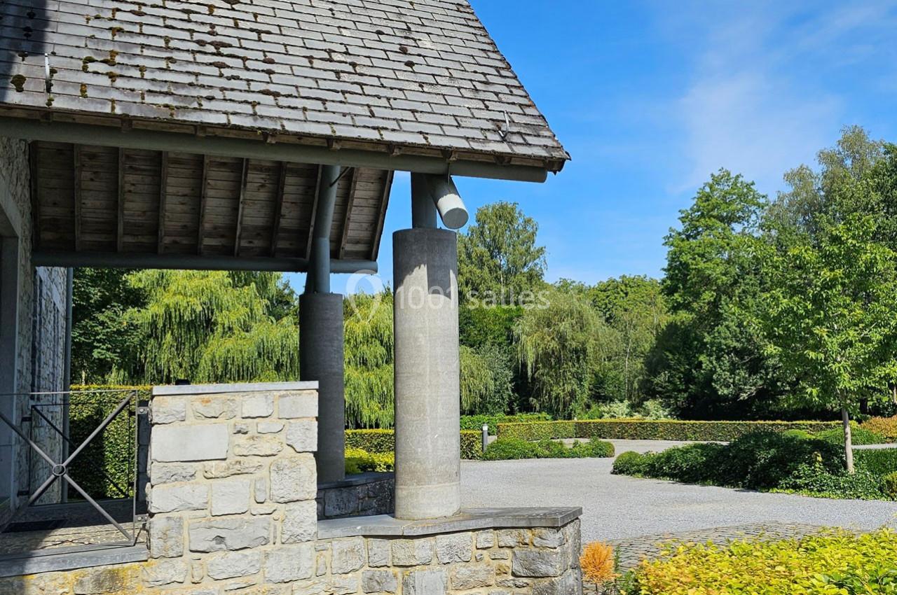 Porche en pierre avec toit en ardoise, colonnes cylindriques et jardin verdoyant en arrière-plan.