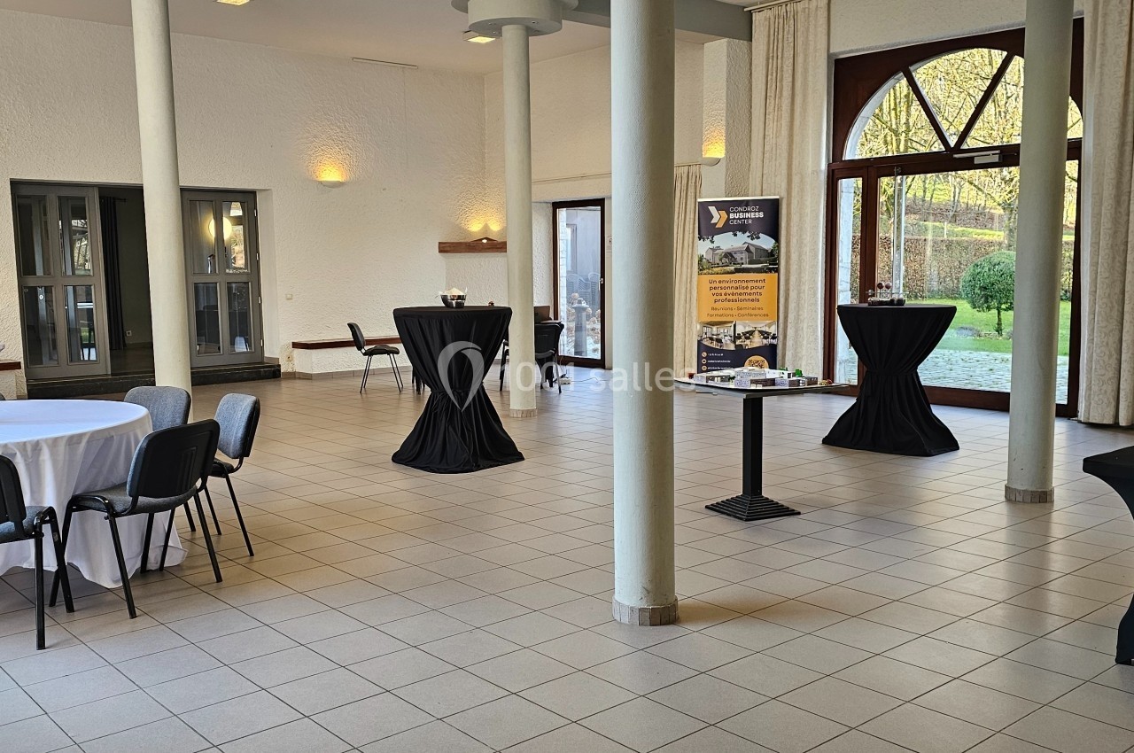 Salle lumineuse avec tables rondes et hautes, nappées de noir et blanc, donnant sur un jardin par de grandes baies vitrées.