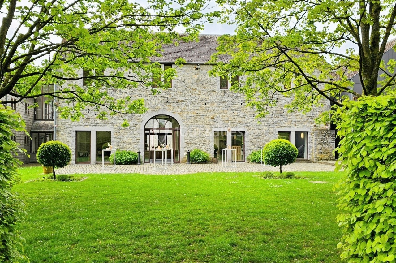 Façade en pierre d'une maison entourée d'arbres et d'arbustes, avec une pelouse verte au premier plan.