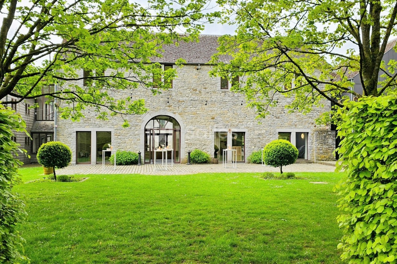 Façade en pierre d'une maison entourée d'arbres et d'arbustes, avec une pelouse verte au premier plan.