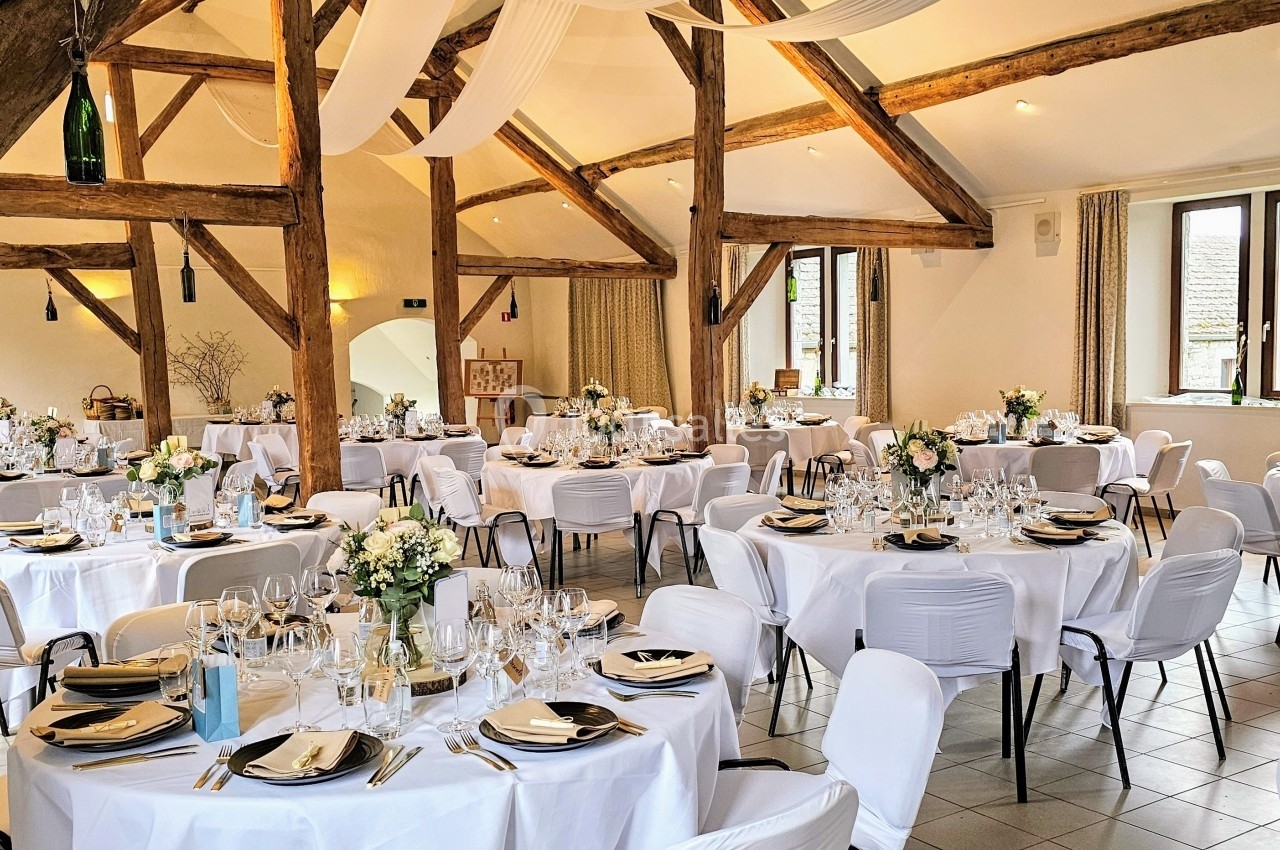 Salle de réception lumineuse avec poutres apparentes, tables rondes dressées avec nappes blanches et décorations florales.