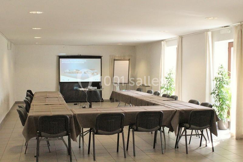 Salle de réunion lumineuse avec tables disposées en U, écran de projection, paperboard et plantes décoratives.
