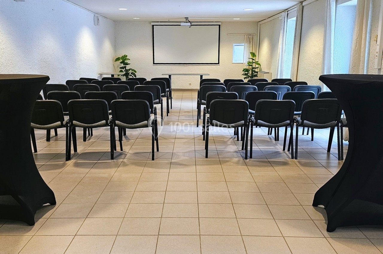 Salle de conférence lumineuse avec rangées de chaises, tables hautes noires et écran de projection au fond.