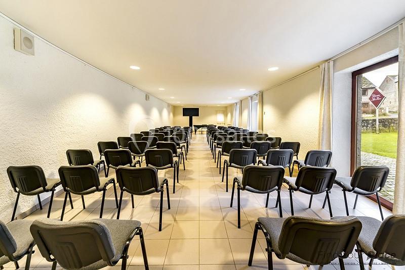 Salle de conférence lumineuse avec rangées de chaises noires alignées, écran au fond et grande baie vitrée à droite.