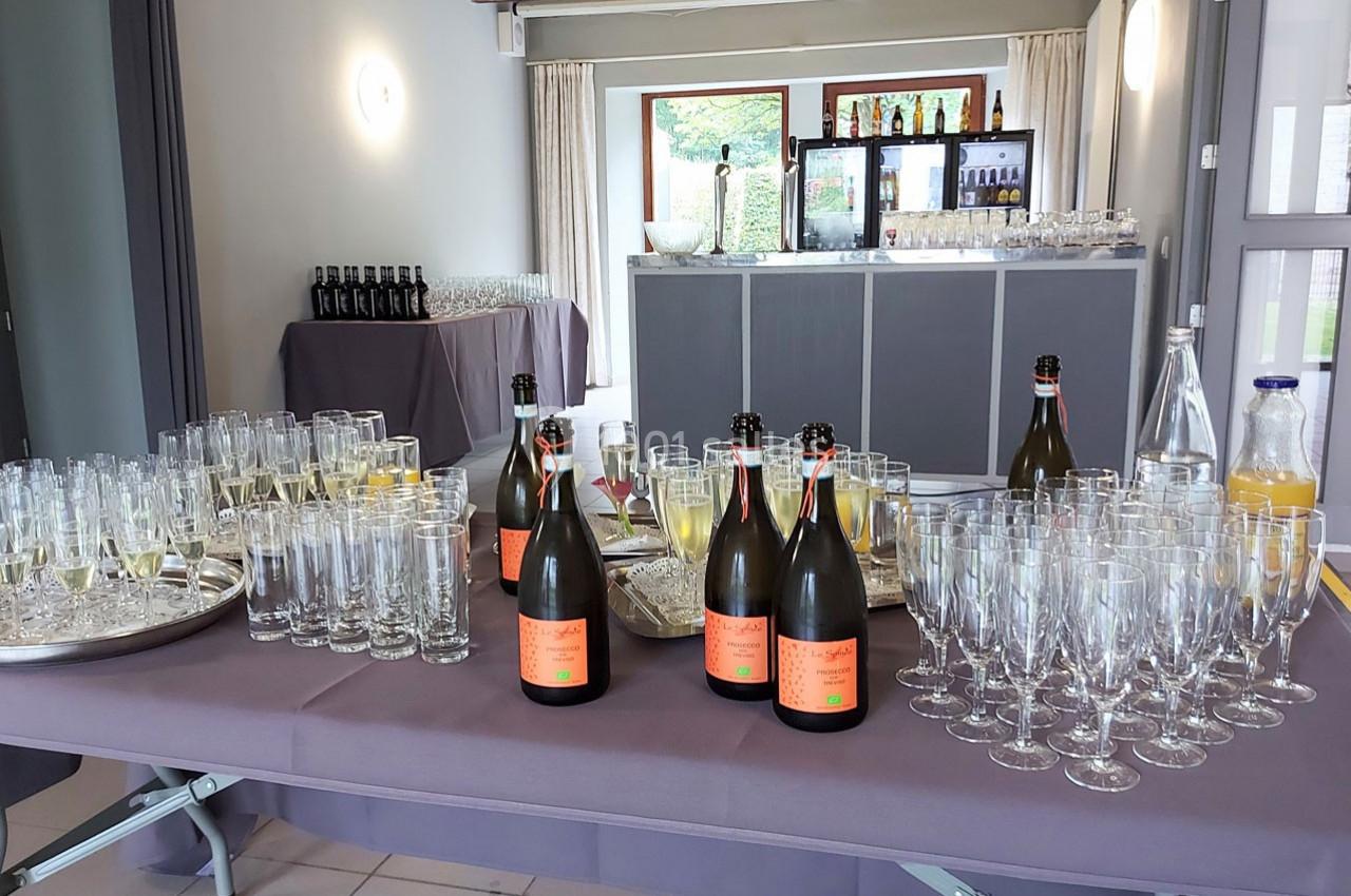 Table dressée avec des bouteilles de champagne, verres vides et remplis, et carafes de jus dans une salle lumineuse.