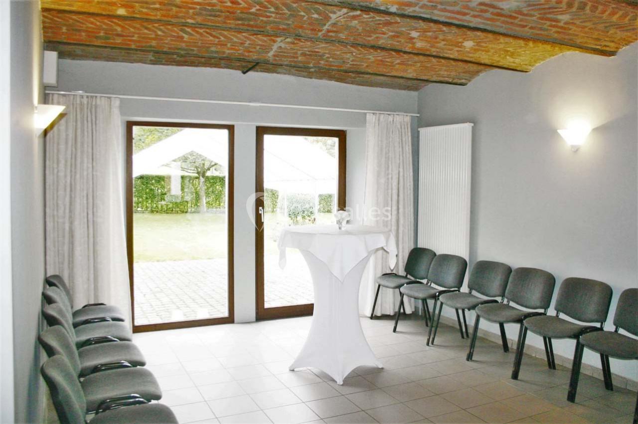 Salle lumineuse avec plafond en briques, chaises alignées contre les murs et table haute près d'une baie vitrée donnant sur…