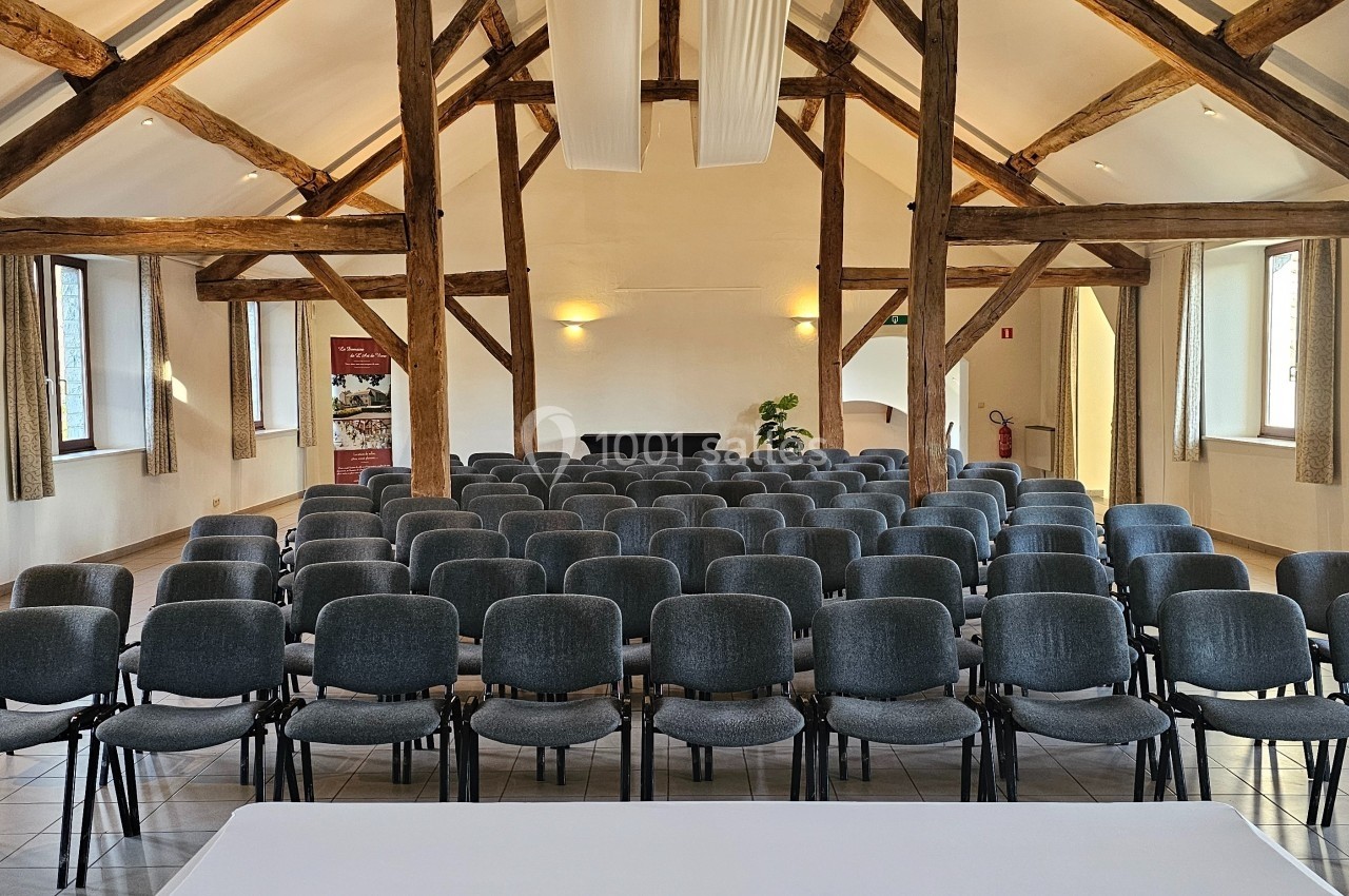 Salle de conférence lumineuse avec des rangées de chaises grises alignées sous une charpente en bois apparente.