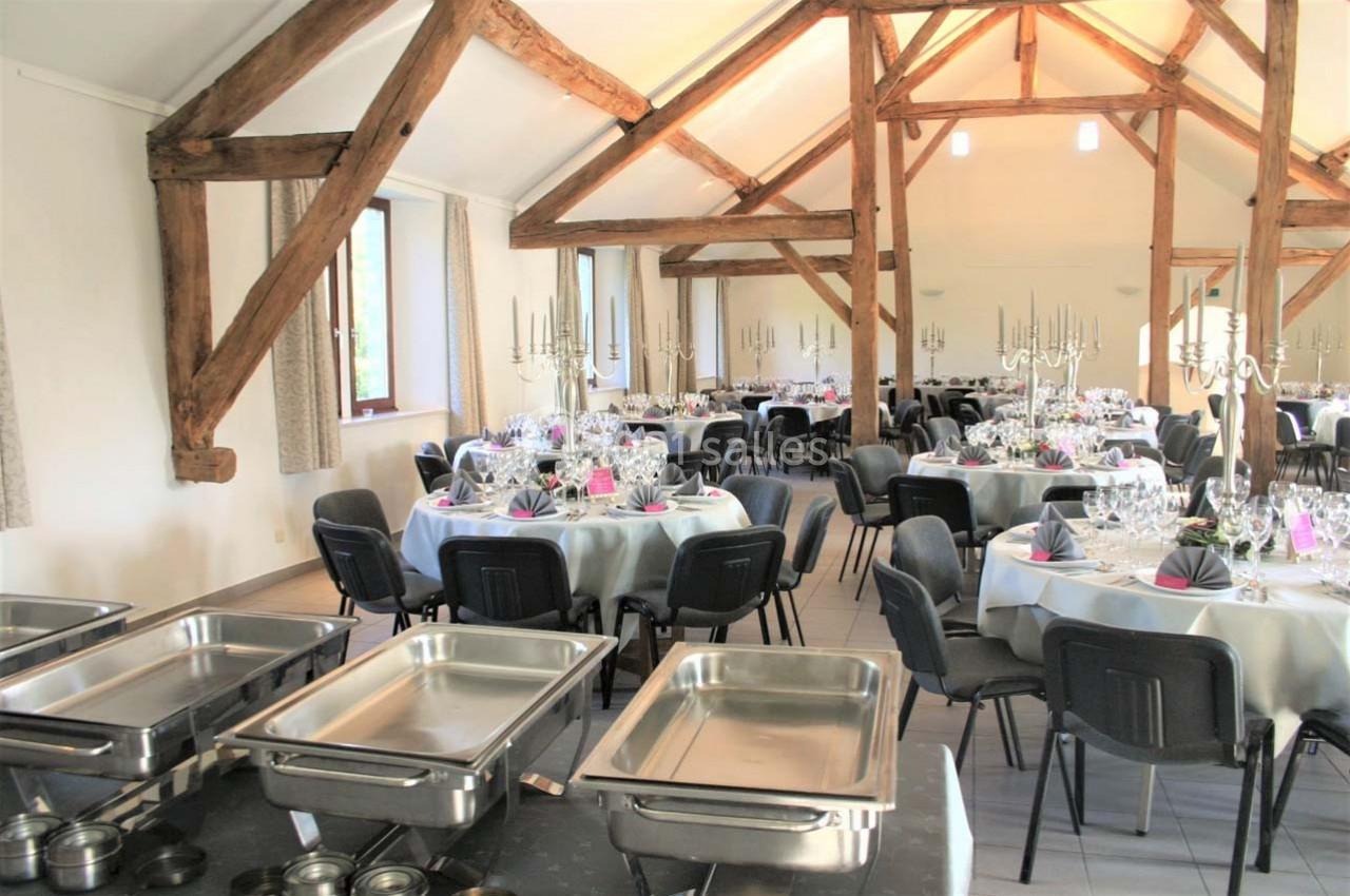Salle de réception lumineuse avec poutres apparentes, tables dressées pour un événement et chafing dishes au premier plan.