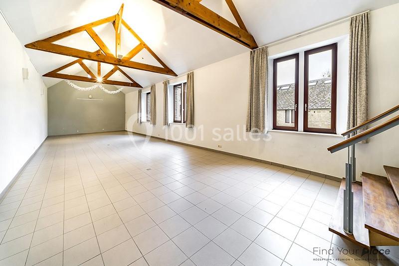 Grande pièce lumineuse avec poutres apparentes, carrelage au sol, fenêtres à rideaux et escalier en bois.