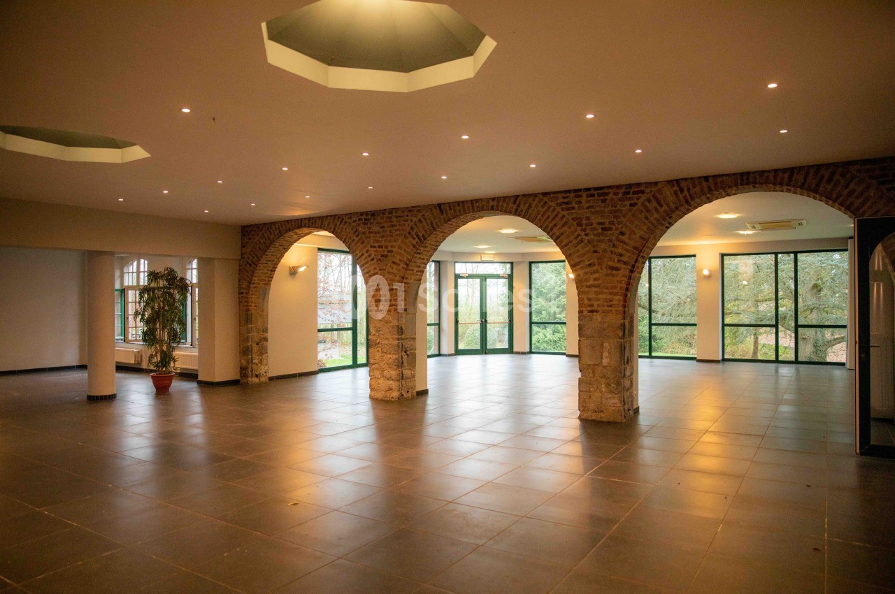 Salle spacieuse avec arches en briques, grandes fenêtres donnant sur un espace vert et sol en carrelage sombre.