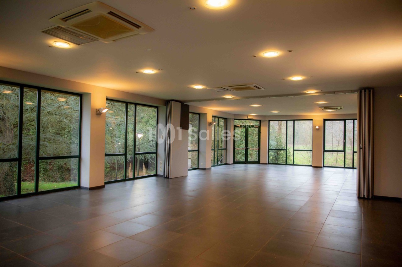 Salle vide avec de grandes baies vitrées donnant sur un espace extérieur arboré, éclairée par des plafonniers.