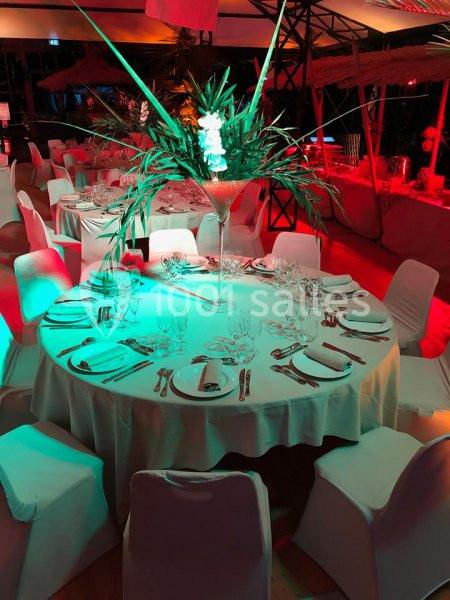 Salle de réception décorée avec des tables rondes dressées, centre de table floral et éclairage coloré.