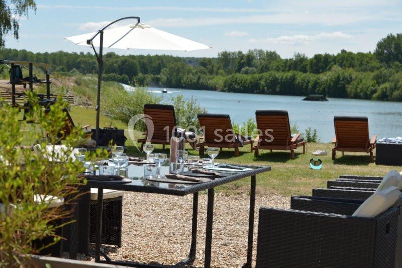 Mobilier de jardin avec table dressée et transats face à une rivière entourée de verdure, par une journée ensoleillée.