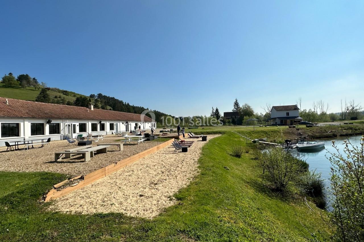 Bâtiments blancs avec toit en tuiles rouges près d'un étang, entourés de verdure sous un ciel dégagé.