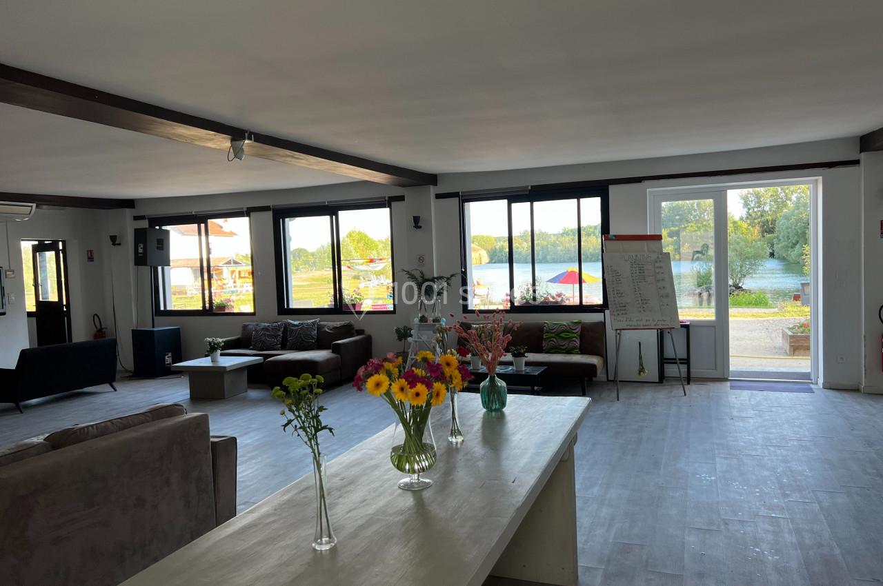 Salle lumineuse avec grandes fenêtres, mobilier moderne, fleurs sur une table et vue sur un espace extérieur verdoyant.