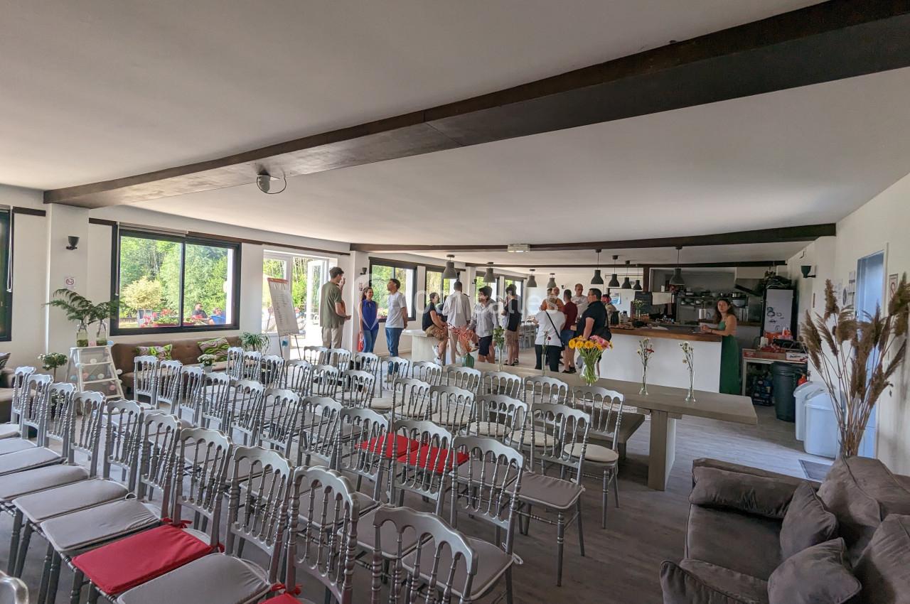 Salle lumineuse avec chaises alignées, un groupe de personnes discute près d'un bar au fond de la pièce.