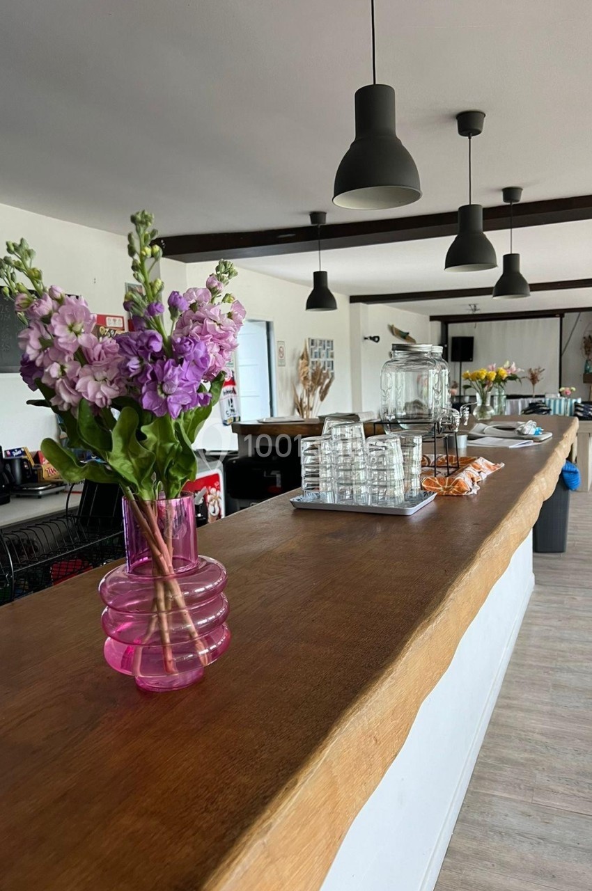 Comptoir en bois avec vase de fleurs violettes au premier plan, éclairage suspendu et espace de restauration en arrière-plan.