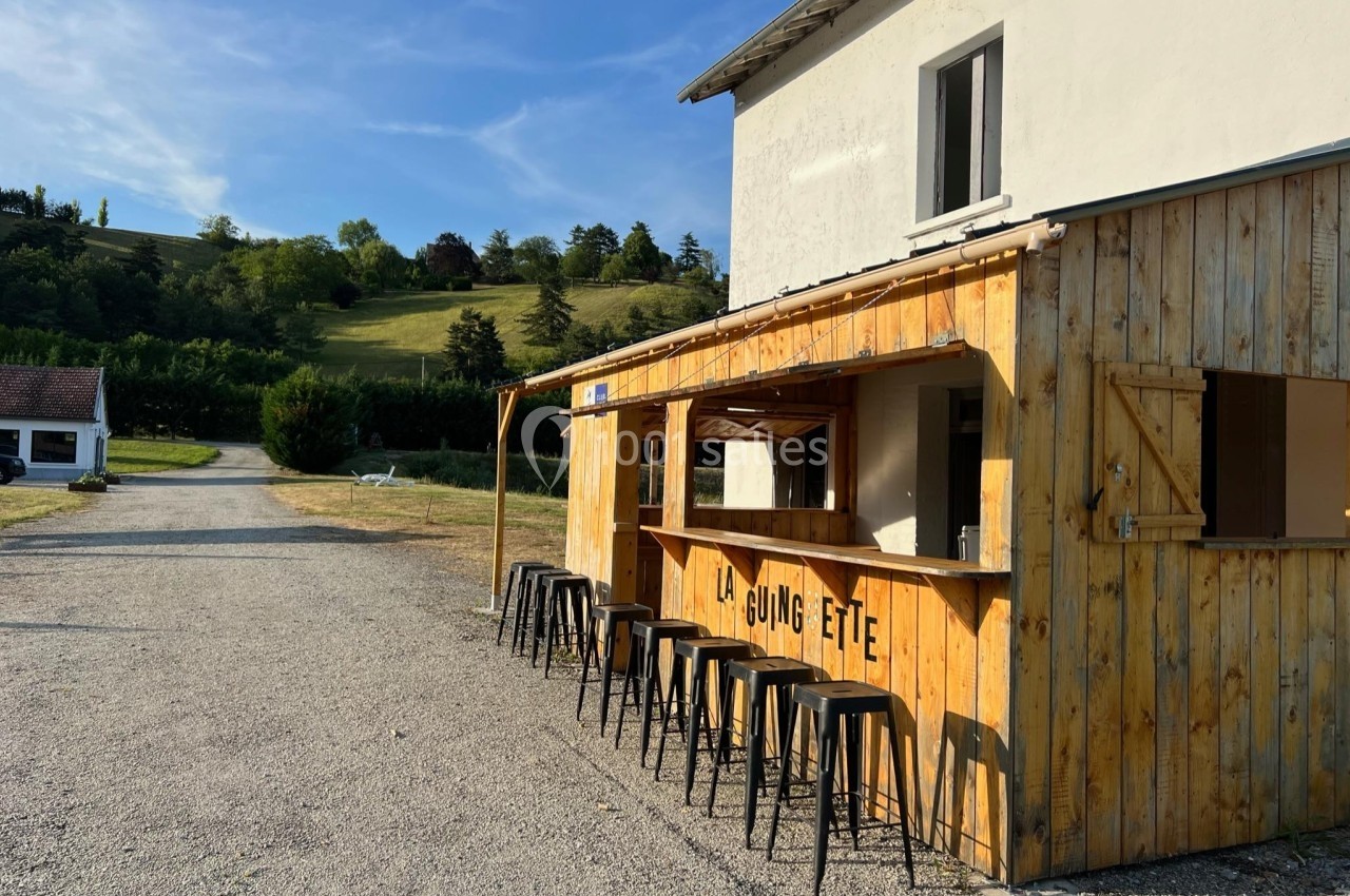 Bar en bois avec tabourets noirs en extérieur, situé près d'un chemin et entouré de collines verdoyantes.