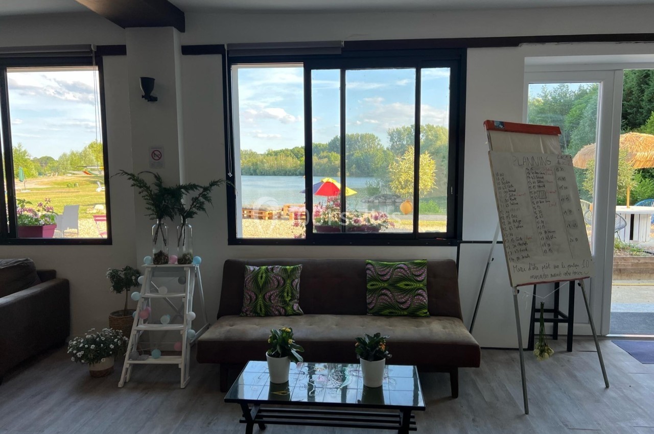 Salon lumineux avec canapé, table basse, plantes, tableau de notes et vue sur un lac à travers de grandes fenêtres.