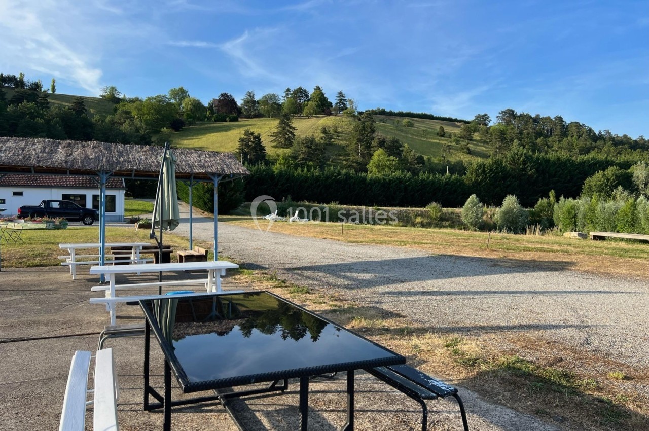 Tables et bancs en plein air sur une terrasse, avec vue sur une colline verdoyante et un ciel dégagé.