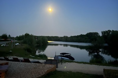 Vue d'une rivière bordée de végétation avec une maison au toit rouge et un ciel partiellement nuageux au coucher du soleil.