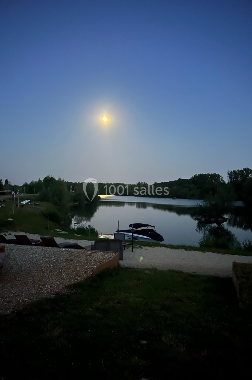 Vue nocturne d'un lac calme éclairé par la lune, entouré de végétation et de quelques bateaux amarrés.