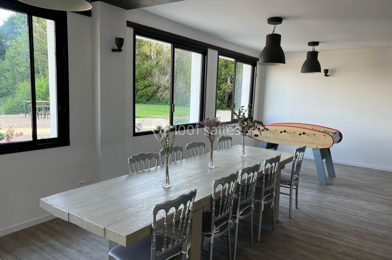 Salle lumineuse avec grande table en bois, chaises grises, baby-foot et fenêtres donnant sur un espace vert.