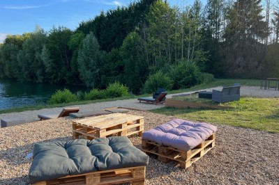 Vue d'une rivière bordée de végétation avec une maison au toit rouge et un ciel partiellement nuageux au coucher du soleil.