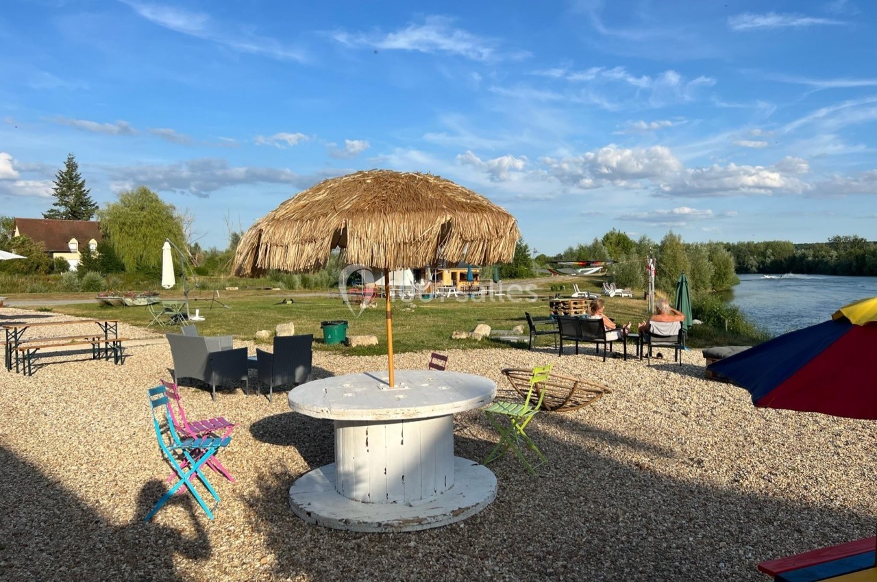 Espace extérieur avec tables, chaises colorées, parasol en paille et vue sur un cours d'eau sous un ciel dégagé.