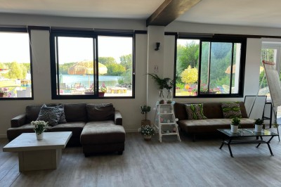 Vue d'une rivière bordée de végétation avec une maison au toit rouge et un ciel partiellement nuageux au coucher du soleil.