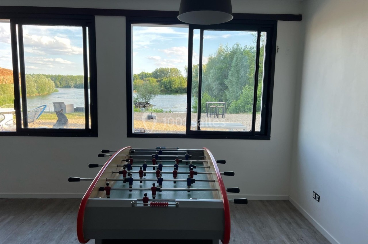 Table de baby-foot dans une pièce lumineuse avec vue sur un paysage de rivière et de verdure à travers de grandes fenêtres.