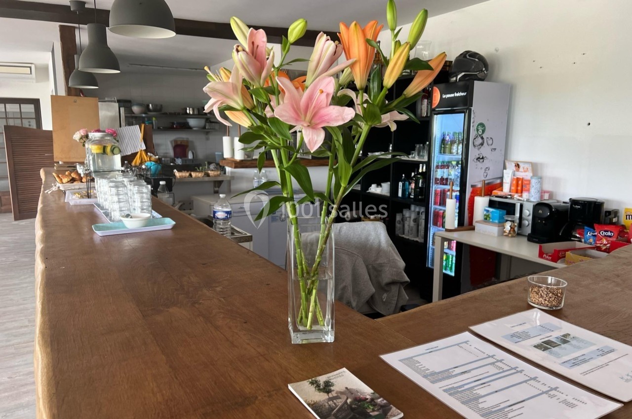 Vase de lys roses et orange posé sur un comptoir en bois dans un espace de restauration avec des équipements en arrière-plan.