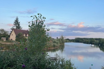 Vue d'une rivière bordée de végétation avec une maison au toit rouge et un ciel partiellement nuageux au coucher du soleil.