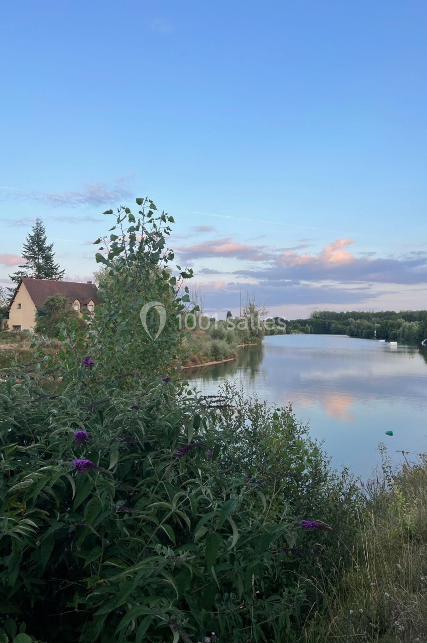 Vue d'une rivière bordée de végétation, avec une maison et un ciel partiellement nuageux au coucher du soleil.