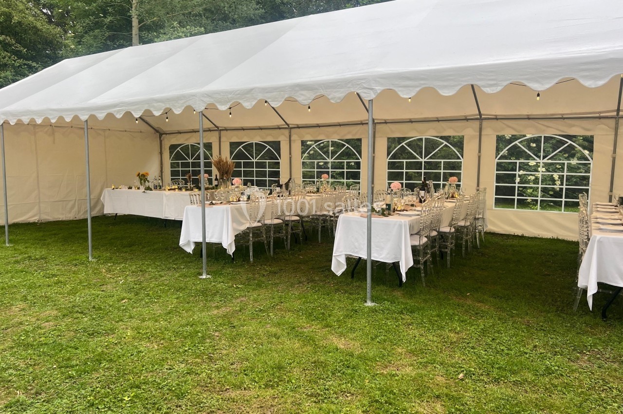Tente blanche installée sur une pelouse, abritant des tables dressées avec des chaises transparentes pour un événement.