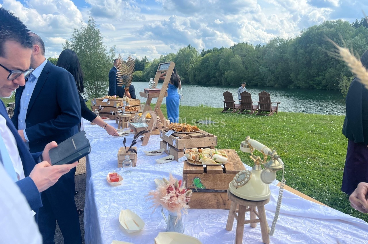 Table de buffet en extérieur près d'un lac, avec des invités en tenue élégante et des chaises disposées au bord de l'eau.