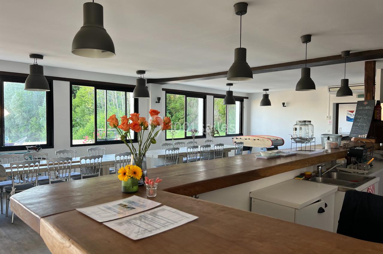 Salle lumineuse avec de grandes fenêtres, un comptoir en bois, des tables dressées et des fleurs en décoration.