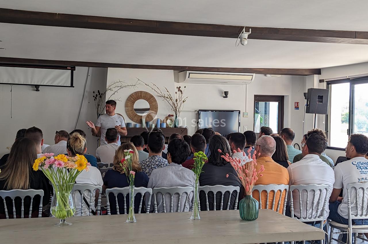 Un groupe de personnes assises écoute un intervenant dans une salle lumineuse avec des décorations florales.
