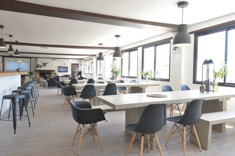 Salle lumineuse avec de grandes fenêtres, tables en bois clair, chaises noires et décoration minimaliste.