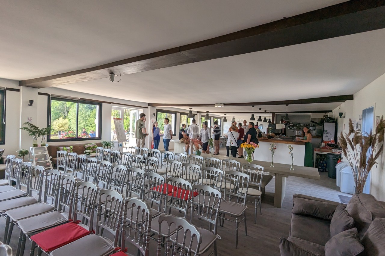 Salle lumineuse avec des rangées de chaises, un bar au fond et des personnes debout discutant près des fenêtres.
