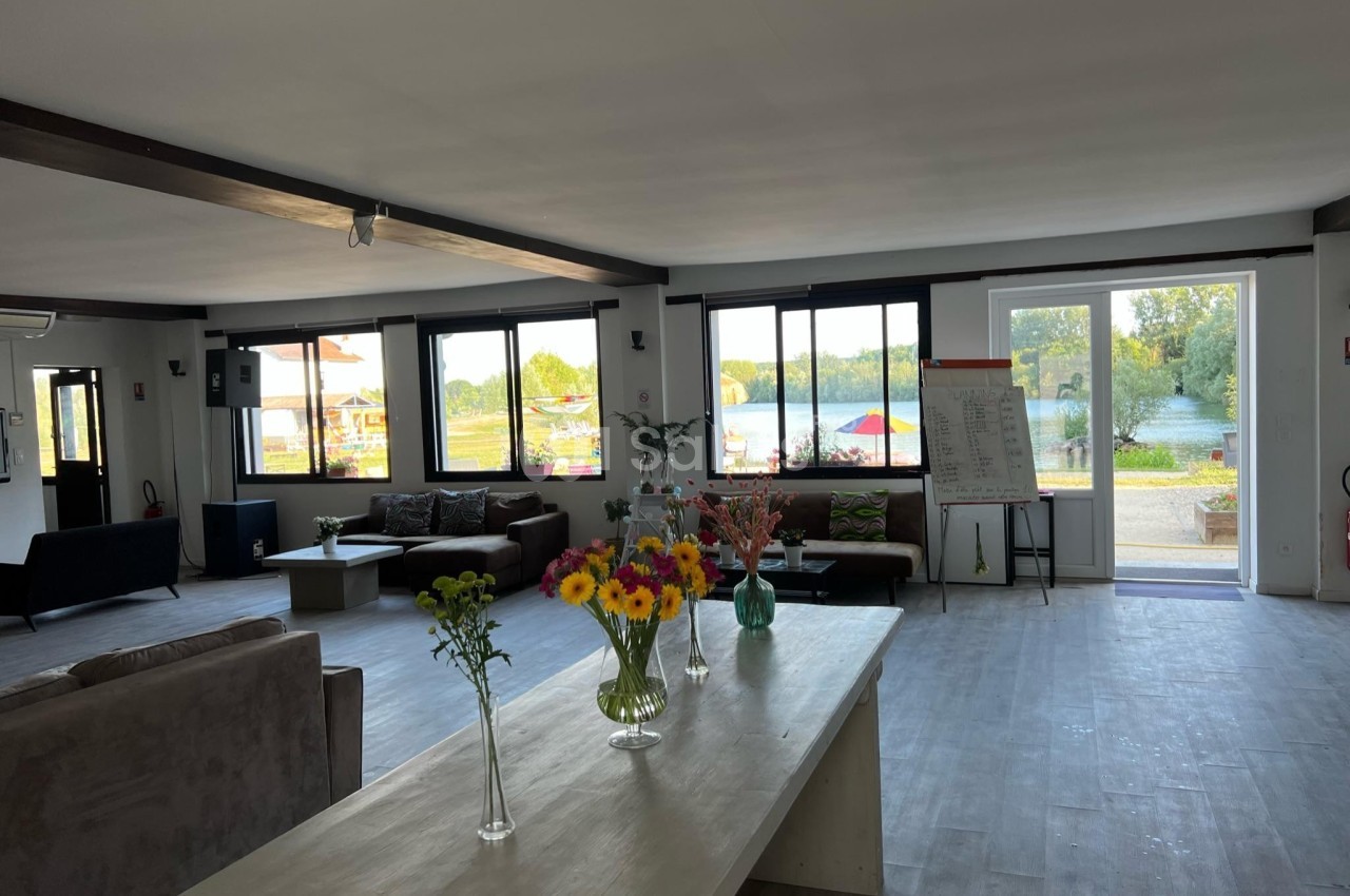 Salle lumineuse avec grandes baies vitrées, mobilier moderne, fleurs sur une table et vue sur un espace extérieur verdoyant.