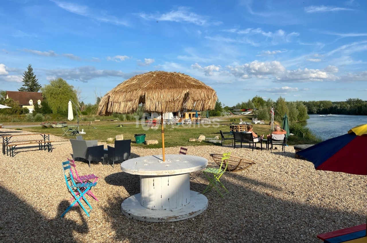 Espace extérieur avec tables, chaises colorées et parasol en paille, près d'une rivière sous un ciel dégagé.