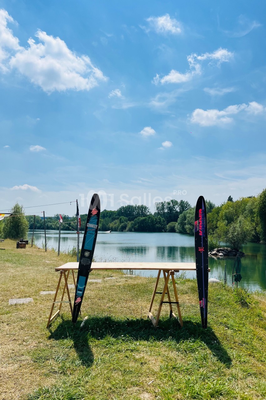 Deux skis nautiques posés contre une table en bois, sur une pelouse près d'un lac entouré de verdure sous un ciel bleu.
