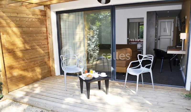 Terrasse en bois avec deux chaises, une table basse et vue sur l'intérieur d'une chambre moderne par une baie vitrée.